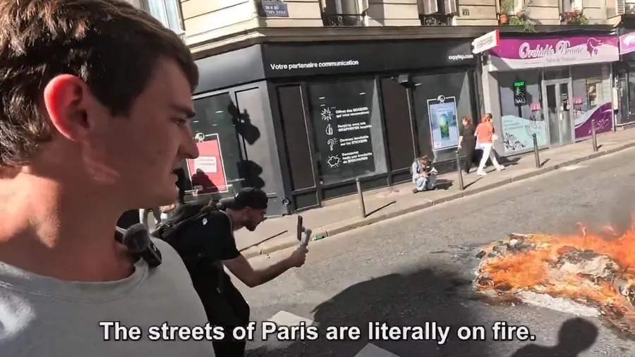 Youtuber Nick Shirley natáča seba uprostred protestu, za ním horí oheň